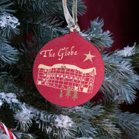 A round red Christmas ornament with gold lettering reading 'The Globe' and a gold illustration of a theatre, hanging on a flocked green Christmas tree with candy canes and a dark red curtain background.