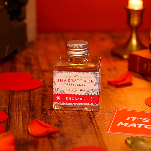 Bottle of Shakespeare Distillery gin on a wooden surface with red flower petals and a candle in the background.