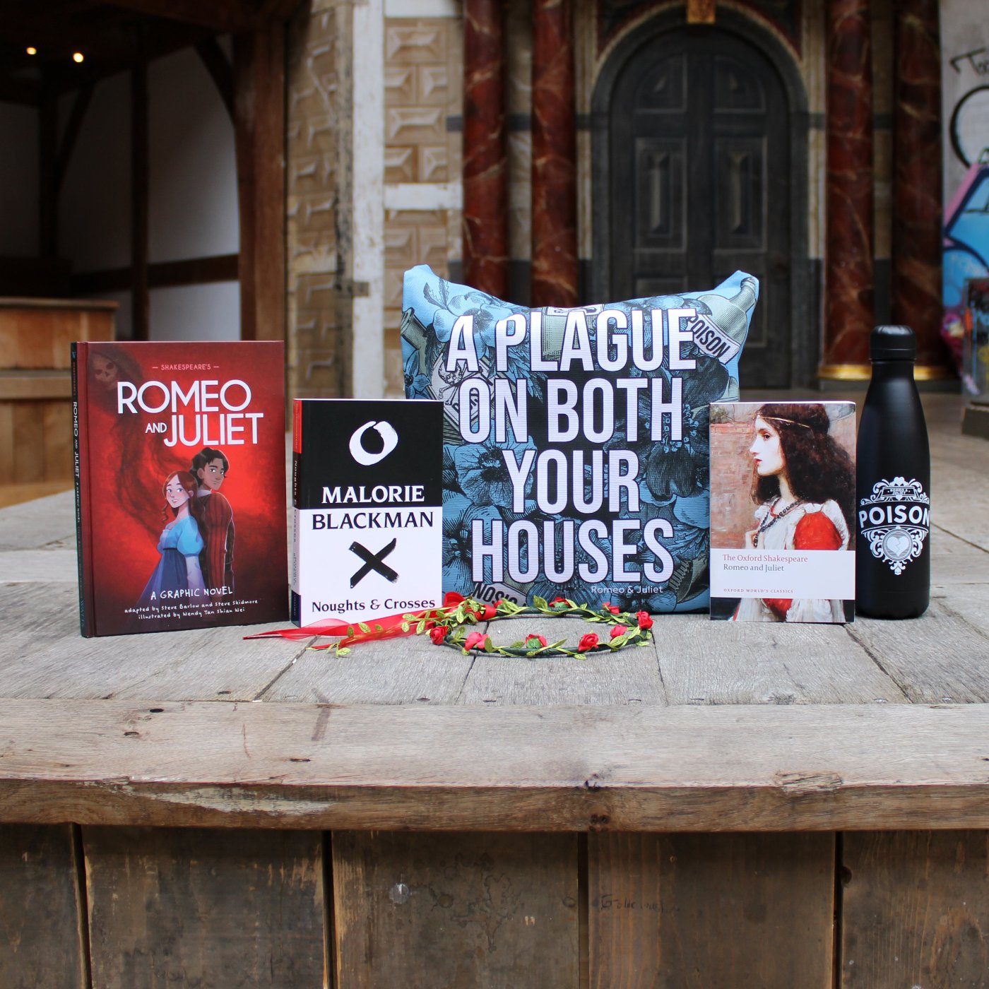 Romeo and Juliet themed gifts on wood panel Globe stage, left to right, red flower crown, hardback book, paperback book, blue tote bag, paperback book, black drinks bottle