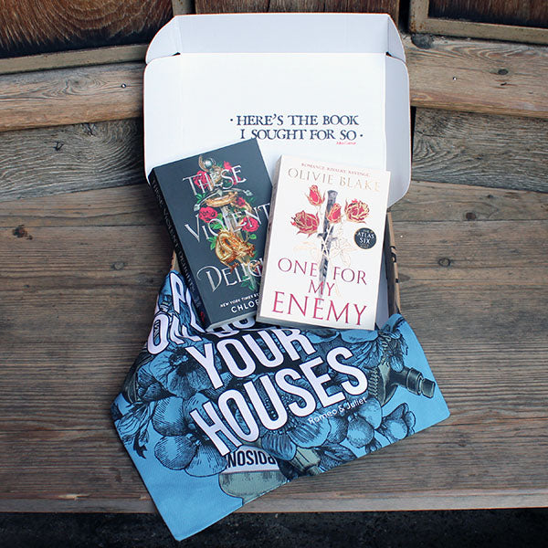 Book box open on wooden step, with black paperback book next to beige paperback book with pale blue patterned cotton tote bag in front