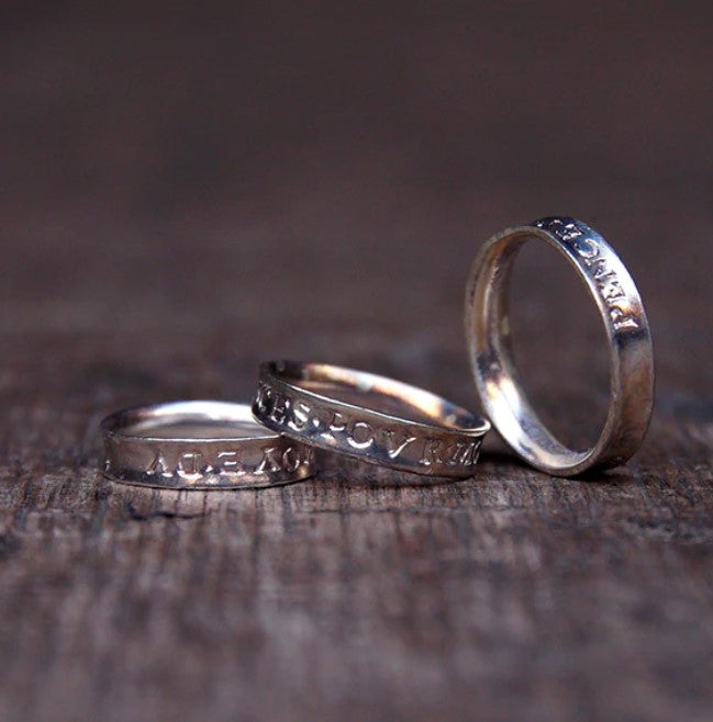 Trio of silver rose rings with Latin text inscribed on them, sitting on grey grainy wood panel
