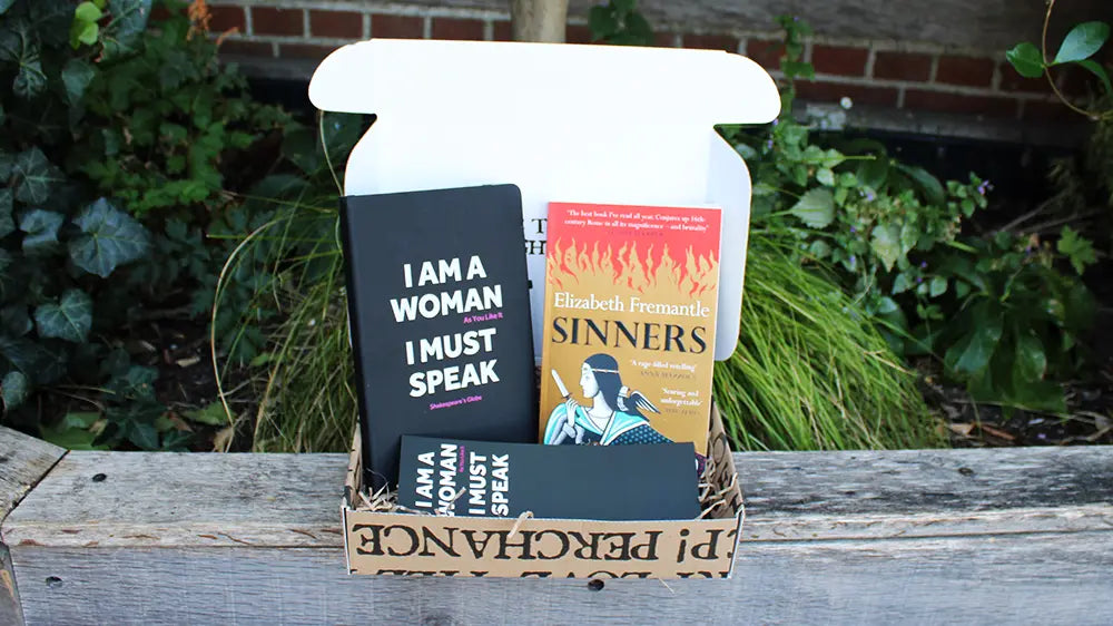 An open cardboard book box sits on a wooden table outdoors, with plants and a brick wall behind it. Inside are Elizabeth Fremantle’s Sinners, an “I Am a Woman, I Must Speak” notebook, and a matching bookmark.