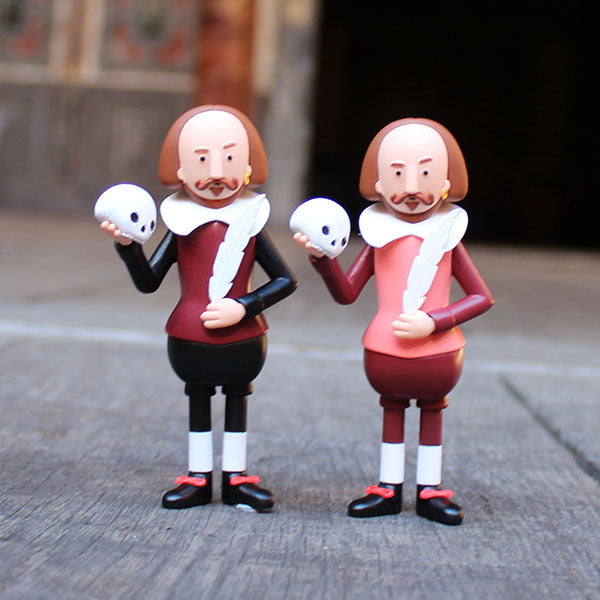 two plastic figures of Shakespeare, one wearing a black and deep red costume and the other in a pink and deep red costume, both holding a white skull and feather