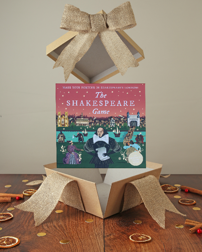 Green and pink square box with cartoon Shakespeare on the cover, popping out of kraft box with bow on wood base with Christmas decor