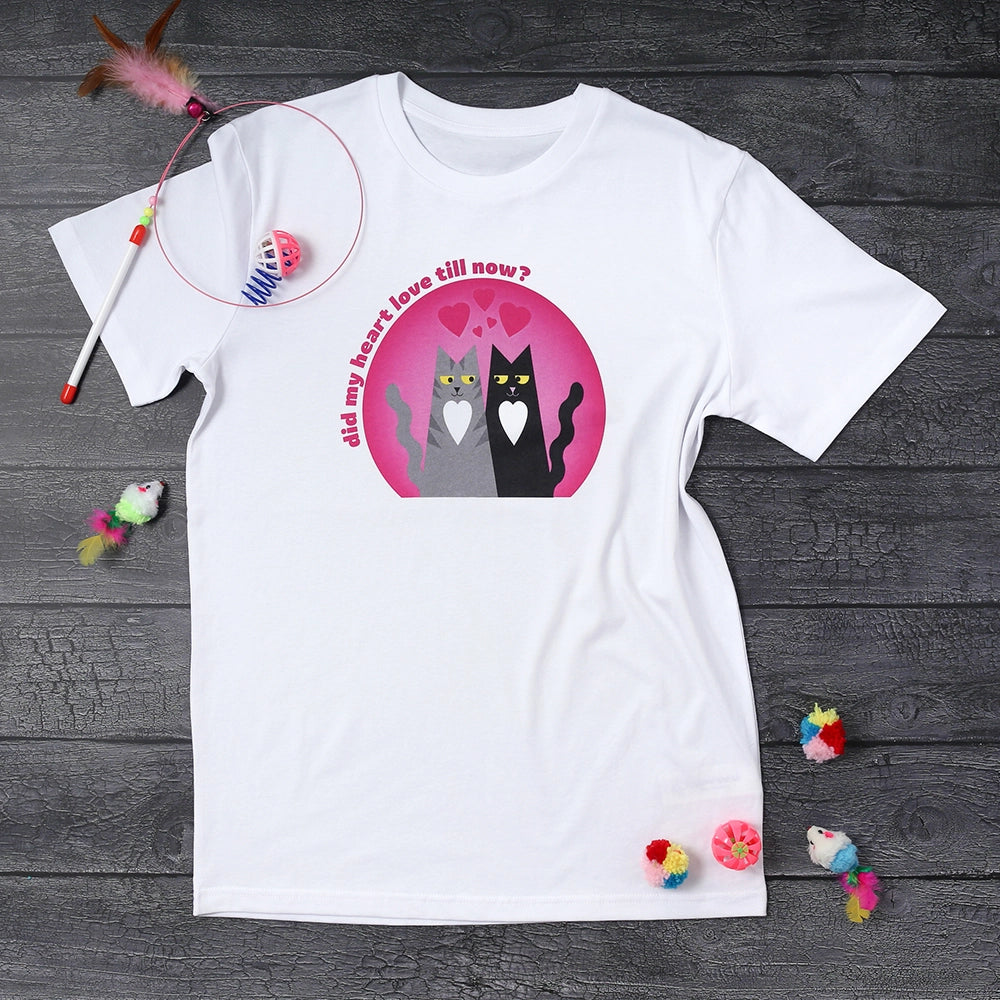 white t-shirt printed with a pink circle in which sit a grey tabby cat and a black cat on a wooden background surrounded by cat toys