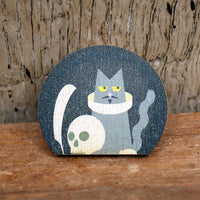Wooden lapel pin featuring an illustrated grey cat with yellow eyes, wearing a white ruff collar, alongside a skull and quill pen, inspired by Shakespeare, displayed on a wooden surface.