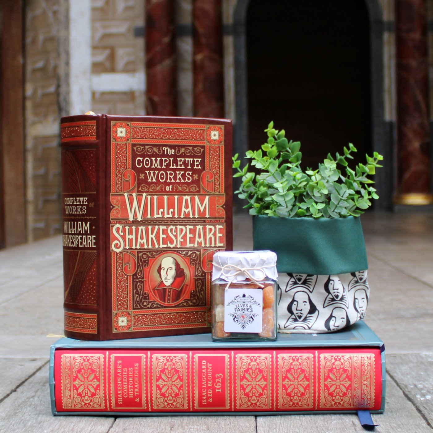 From left to right, brown leatherbound book, black, white, and green printed cloth plant pot, red and blue hardback book, glass jar of sweets on wood stage