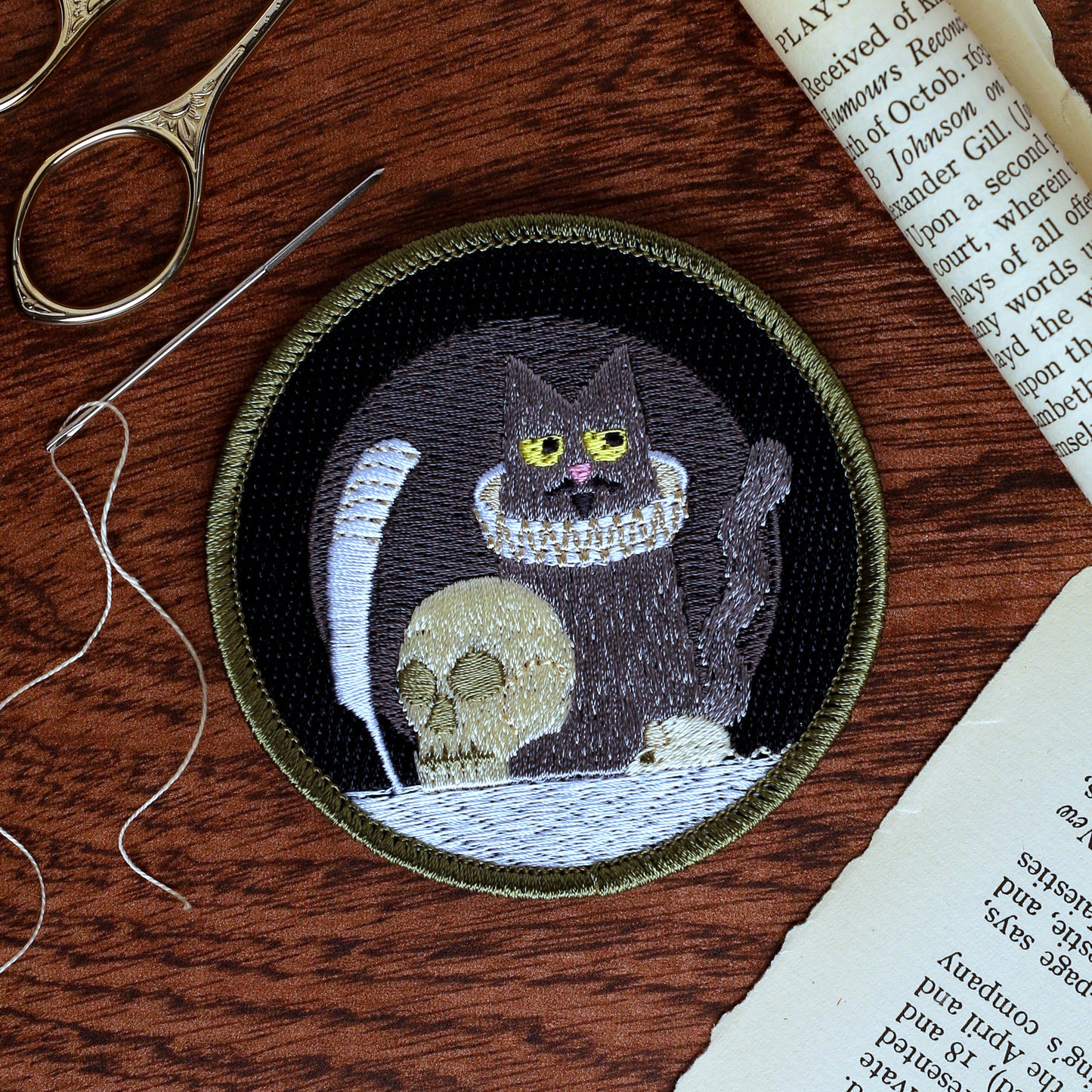 Embroidered patch of a cat with a skull on a wooden surface with sewing tools.