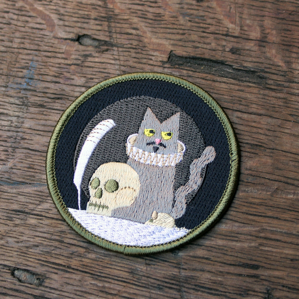 
                      
                        Grey, black and white circular embroidered sew on badge with cat holding a quill in the centre with prop skull, featuring olive green border
                      
                    