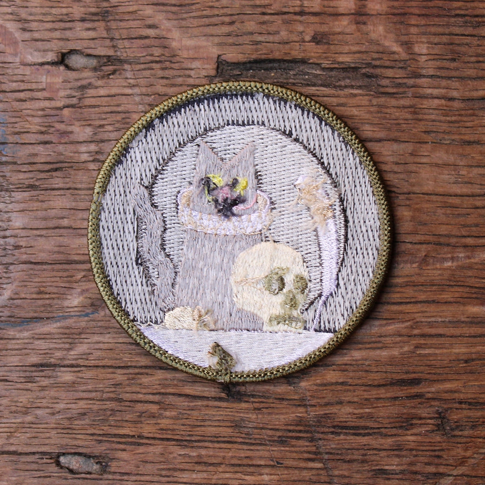 
                      
                        Grey, black and white circular embroidered sew on badge with cat holding a quill in the centre with prop skull, featuring olive green border, from reverse side
                      
                    