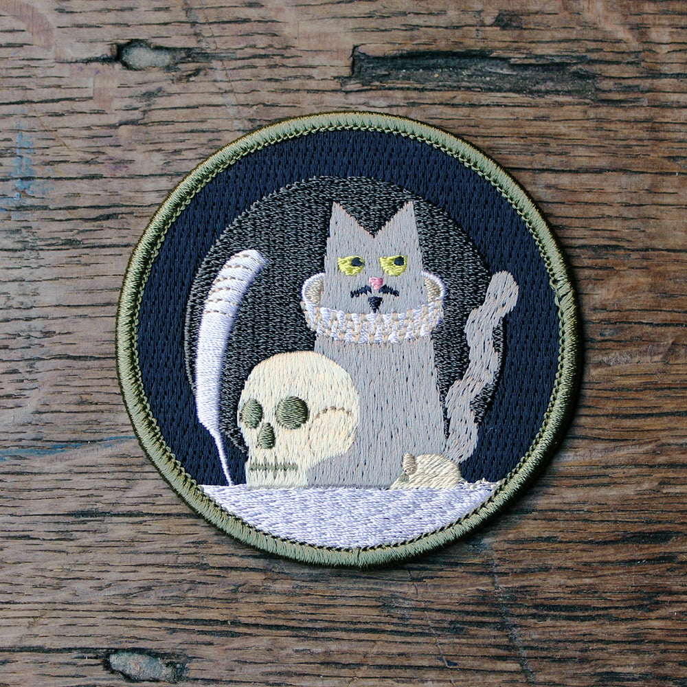 
                      
                        Grey, black and white circular embroidered sew on badge with cat holding a quill in the centre with prop skull, featuring olive green border
                      
                    