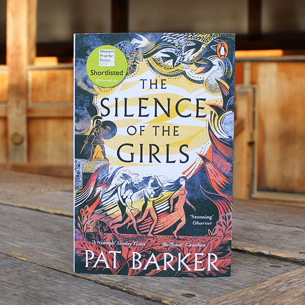 Book titled 'The Silence of the Girls' by Pat Barker on a wooden surface with a blurred background.