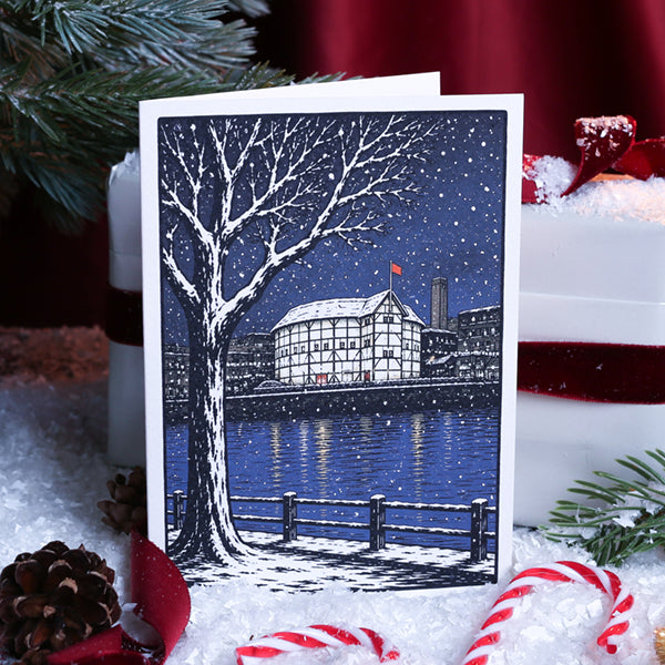 A greetings card featuring a winter cityscape illustration with a snow-covered tree, river, buildings, and falling snow, photographed standing upright a background of Christmas decorations.