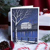 A greetings card featuring a winter cityscape illustration with a snow-covered tree, river, buildings, and falling snow, photographed standing upright a background of Christmas decorations.