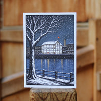 A greetings card featuring a winter cityscape illustration with a snow-covered tree, river, buildings, and falling snow, photographed standing upright against a blurred wooden background.