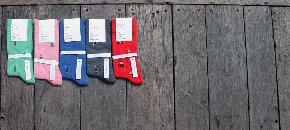 Pairs of 5 Shakespeare themed socks on wood panel stage, from left to right, teal, pink, blue, grey, red