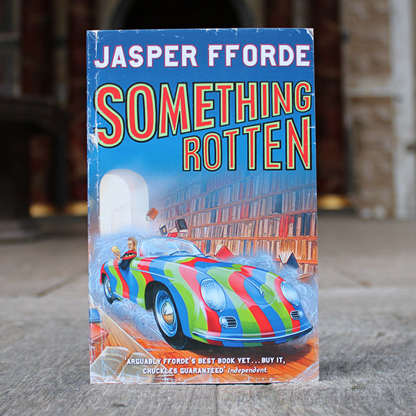 Royal blue paperback book with neon green and red writing and a race car driving through a bookstore on the cover