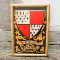 Decorative shield with a coat of arms and 'Sir John St. John' inscription on a wooden background