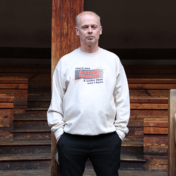 A man wearing a sand coloured sweatshirt with a red and black print. He is standing in front of wooden seating