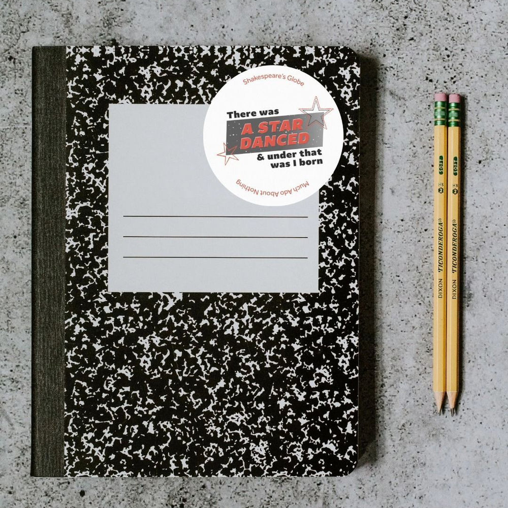 White circular sticker with 1990s style graphic text print, red and black text over gradient of grey and star designs, stuck to retro style notebook sitting on concrete surface with 2 pencils beside it