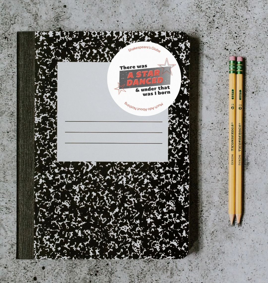 White circular sticker with 1990s style graphic text print, red and black text over gradient of grey and star designs, stuck to retro style notebook sitting on concrete surface with 2 pencils beside it