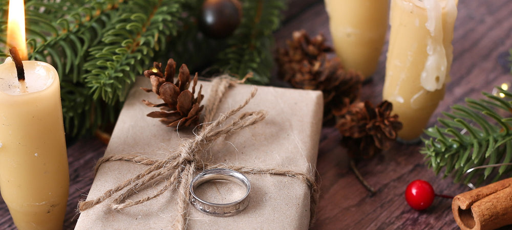 Brown paper package tied with string, on wood panel floor, with natural lit beeswax candles, berries, pinecones and Christmas greenery, featuring a silver ring sitting on the package