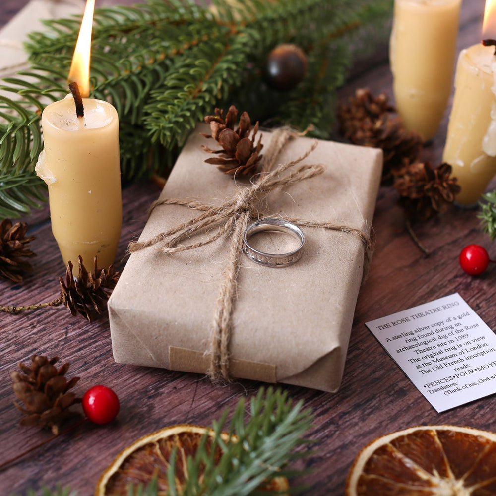 Brown paper package tied with string, on wood panel floor, with natural lit beeswax candles, berries, pinecones and Christmas greenery, featuring a silver ring sitting on the package