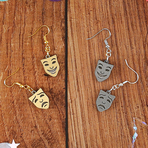 Silver resin mask earrings, comedy and tragedy faces sitting on wooden background, next to gold version of the same earrings