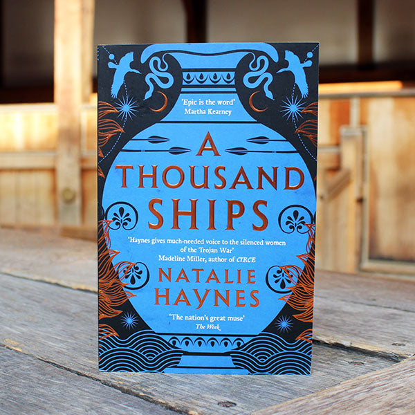 Book cover of 'A Thousand Ships' by Natalie Haynes on a wooden surface.