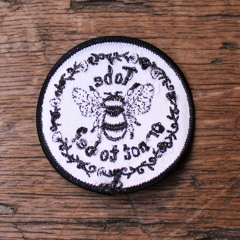 
                      
                        White circular sew on patch with black border and black graphic text and embroidered bee, from reverse side
                      
                    