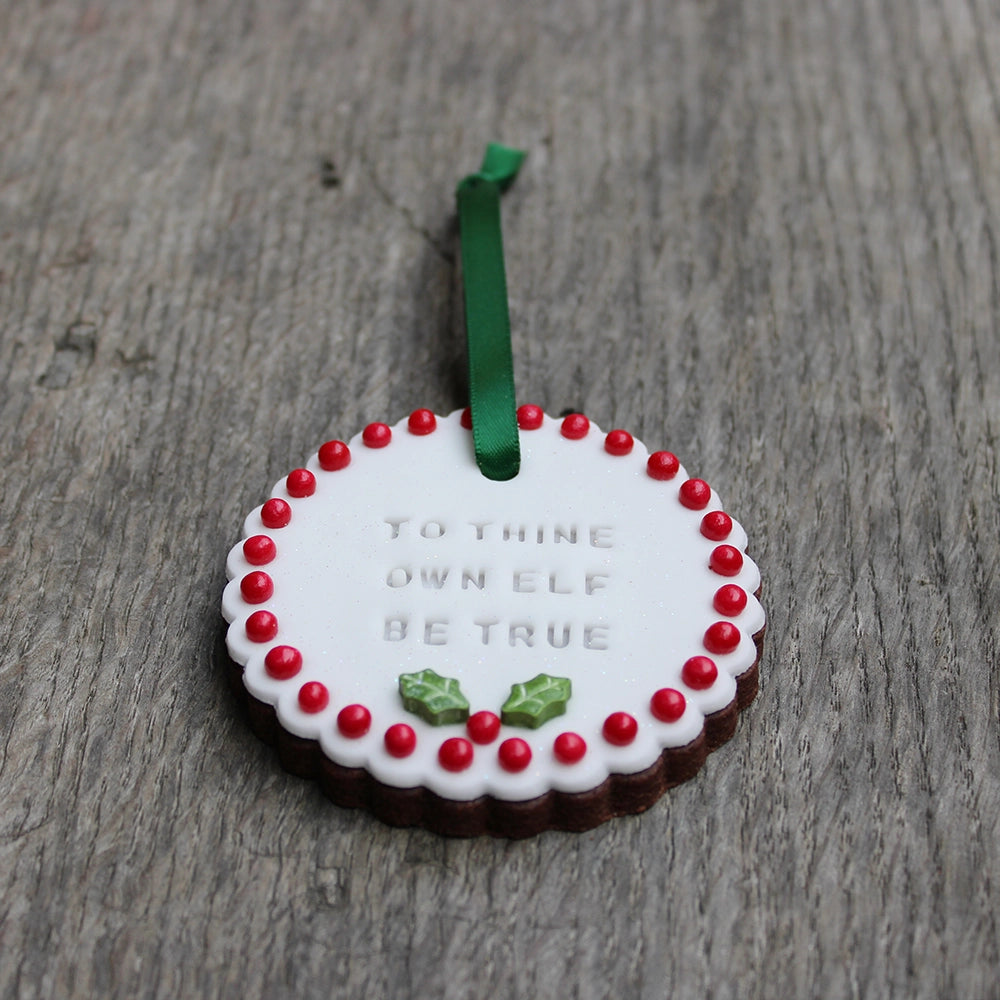 
                  
                    White circular biscuit hanging on forest green ribbon, stamped with text and featuring red ball order and holly leaf at the bottom
                  
                