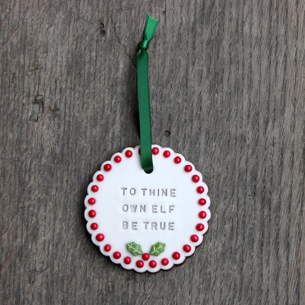 White circular biscuit hanging on forest green ribbon, stamped with text and featuring red ball order and holly leaf at the bottom