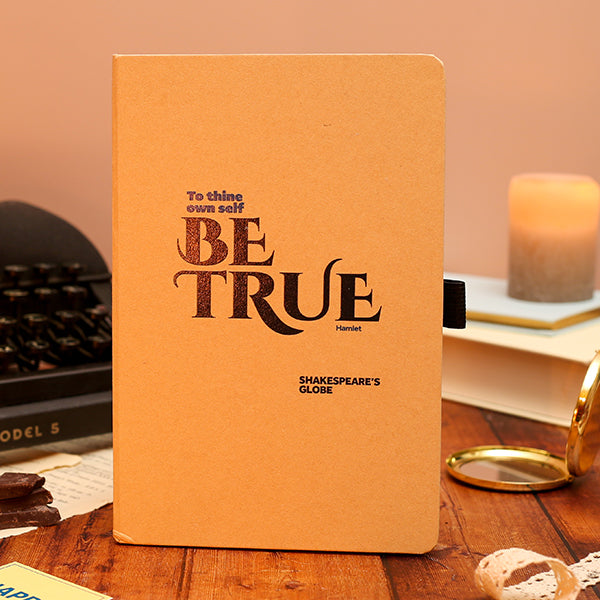 Notebook with 'Be True' quote on a wooden surface with books and a candle in the background