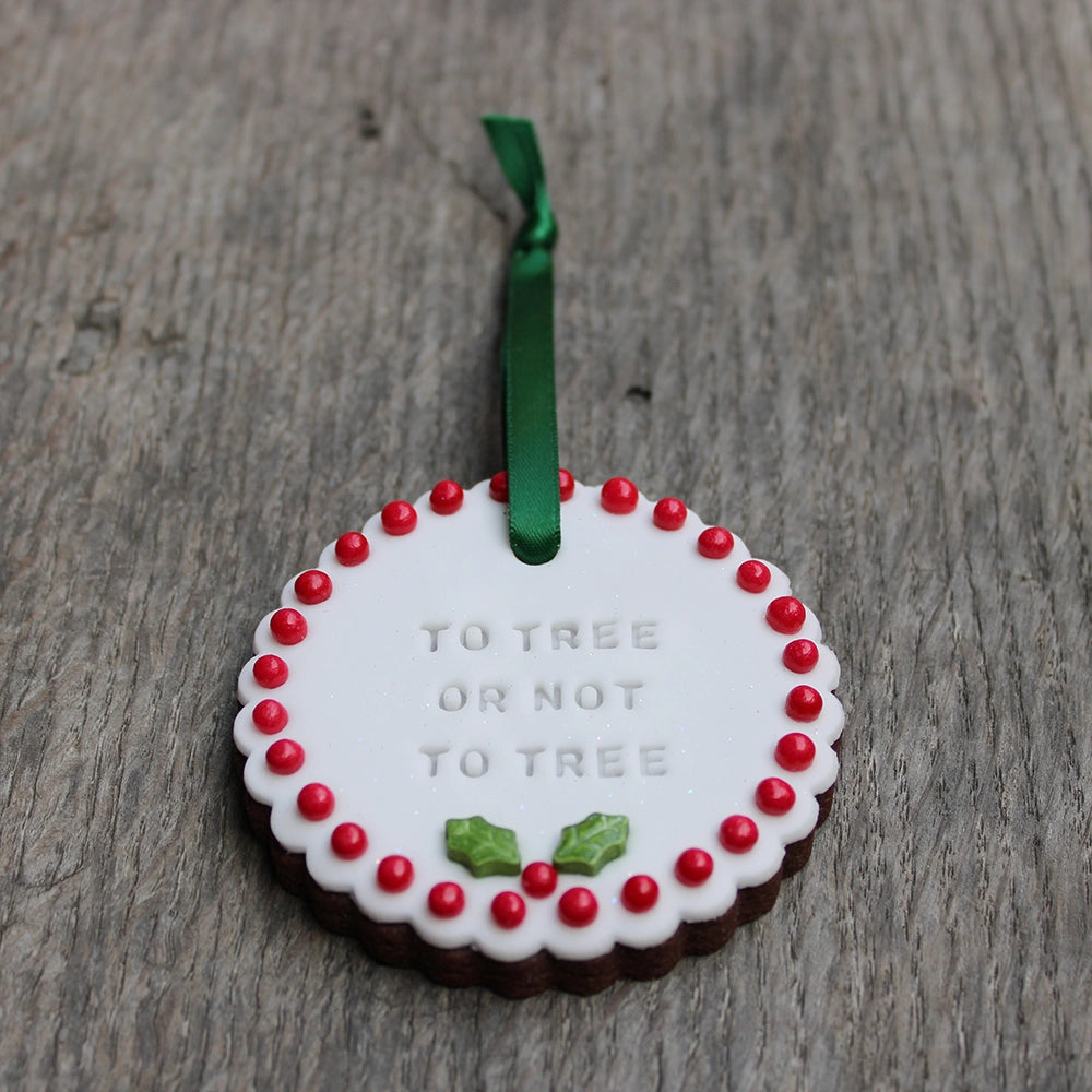 
                  
                    White circular biscuit hanging on forest green ribbon, stamped with text and featuring red ball order and holly leaf at the bottom
                  
                