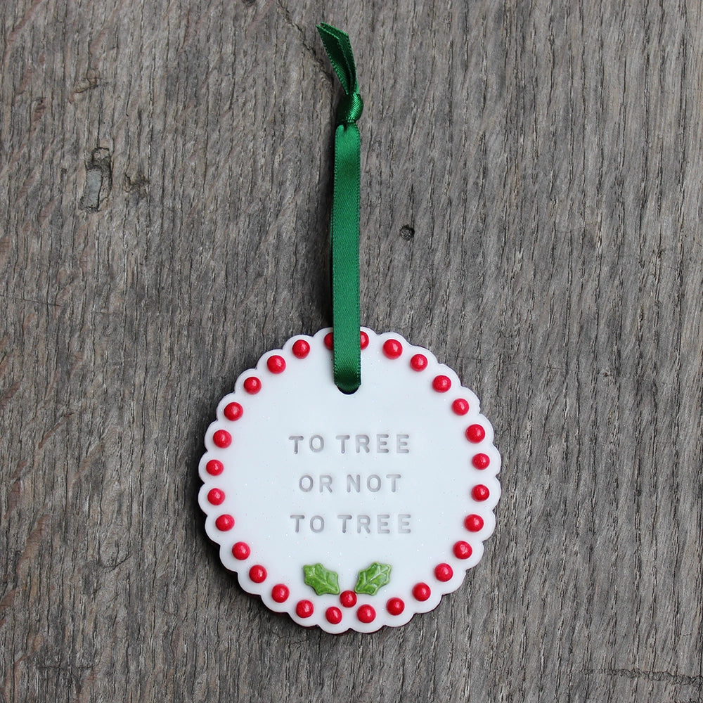 
                  
                    White circular biscuit hanging on forest green ribbon, stamped with text and featuring red ball order and holly leaf at the bottom
                  
                