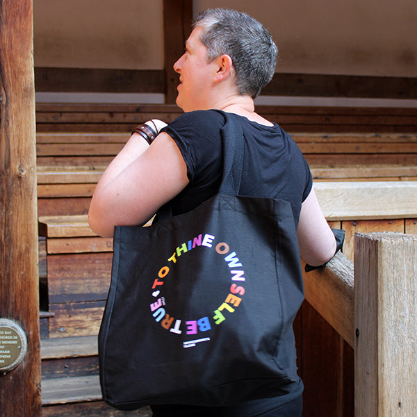 Black cotton tote bag with 2 handles, with circular text graphic showing letters in the colours of the pride flag