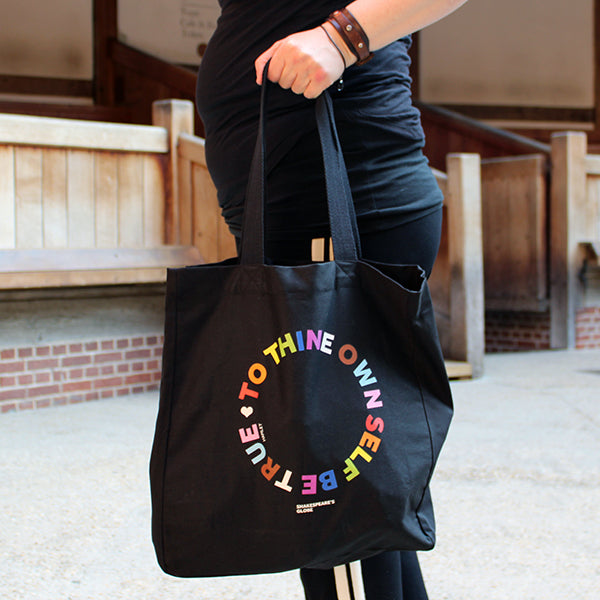 Black cotton tote bag with 2 handles, with circular text graphic showing letters in the colours of the pride flag