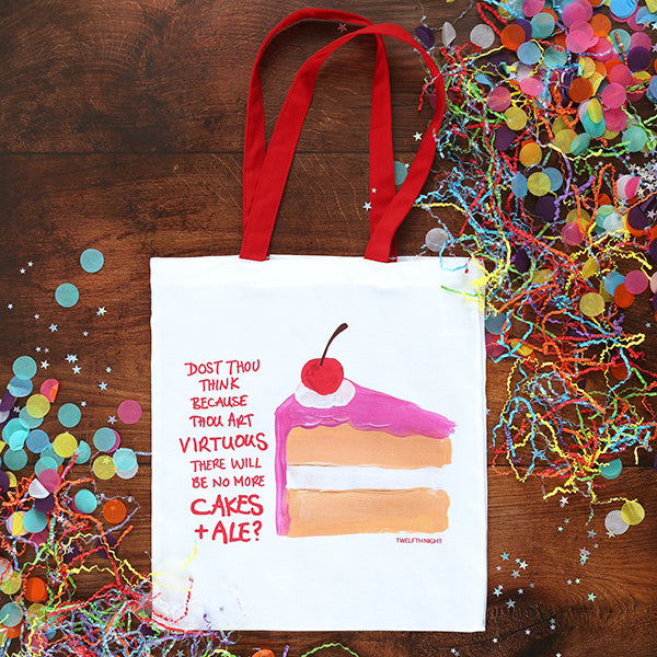 White tote bag with red handles with image of beige cake with pink frosting and red cherry on top, pink graphic text down the left side, on wood floor with confetti scattered