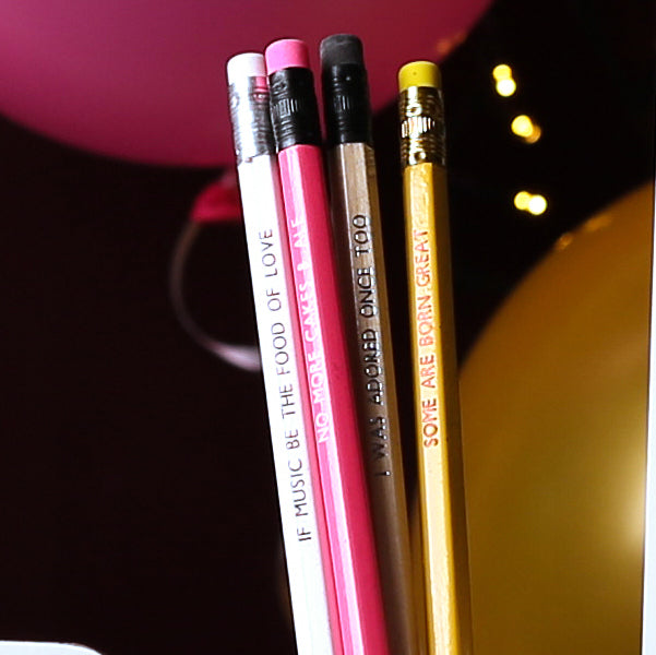 Four quote stamped pencils standing up with erasers to the top of the image, in from of gold and pink balloons on black background, from left to right: white, pink, gold, yellow