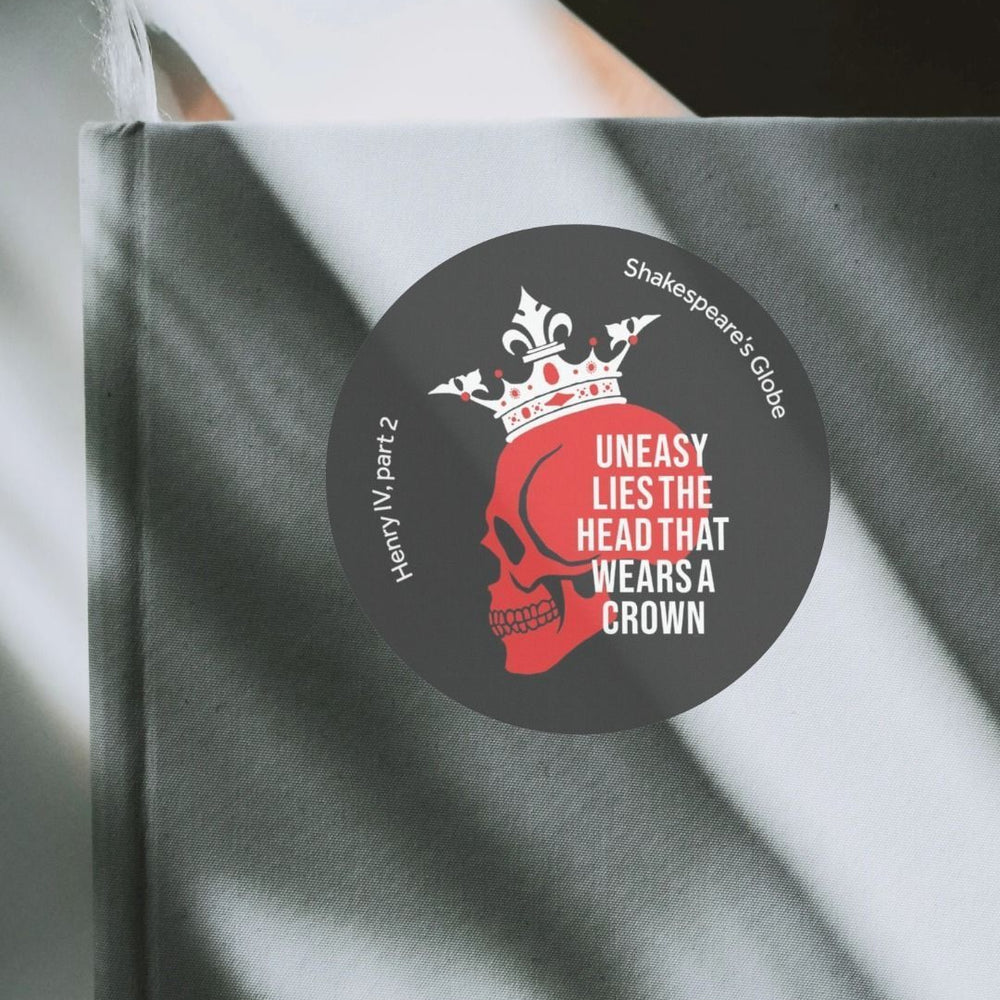 Black circular sticker with red skull wearing white crown, with white text overlayed, on sun streaked grey notebook