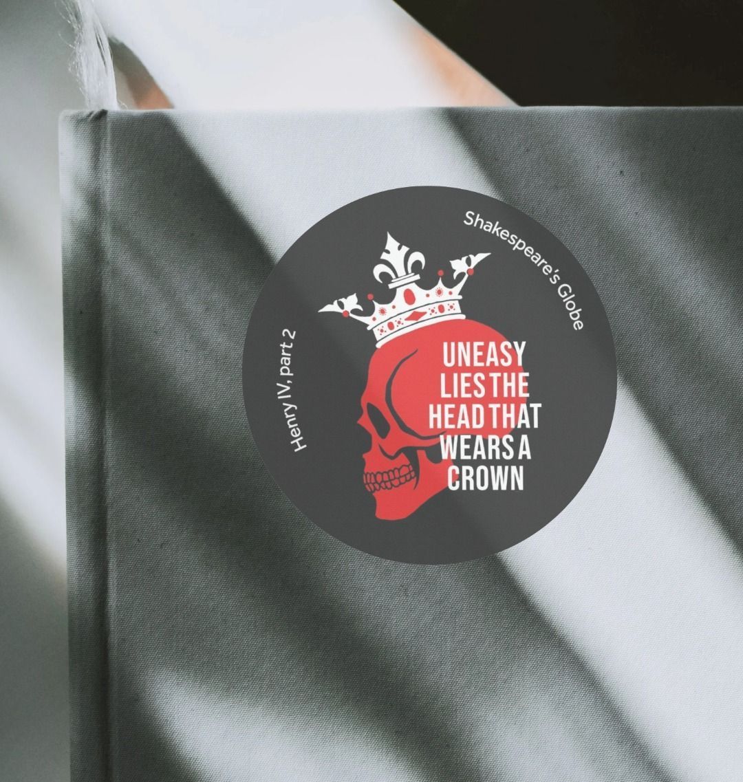 Black circular sticker with red skull wearing white crown, with white text overlayed, on sun streaked grey notebook