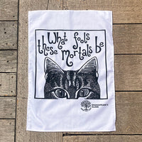 A white tea towel with a black graphic print of a cat peeking up from below and the text 'What fools these mortals be', laid flat on a rustic wooden plank surface. The Shakespeare's Closet logo is printed in the bottom right corner of the design.