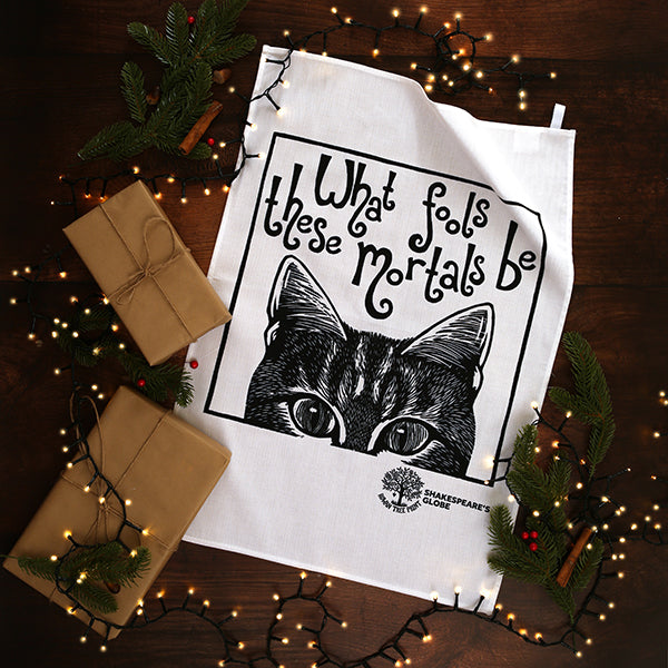 Tea towel with cat illustration and text on a wooden surface with Christmas decorations