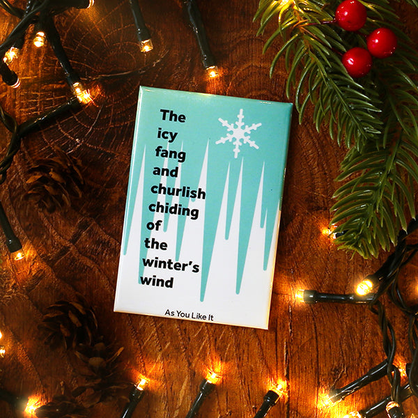 magnet with winter-themed text and snowflake design on a wooden surface with Christmas lights and decorations.