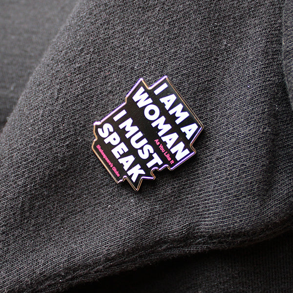 Pin with 'I AM A WOMAN I MUST SPEAK' text on a dark fabric background