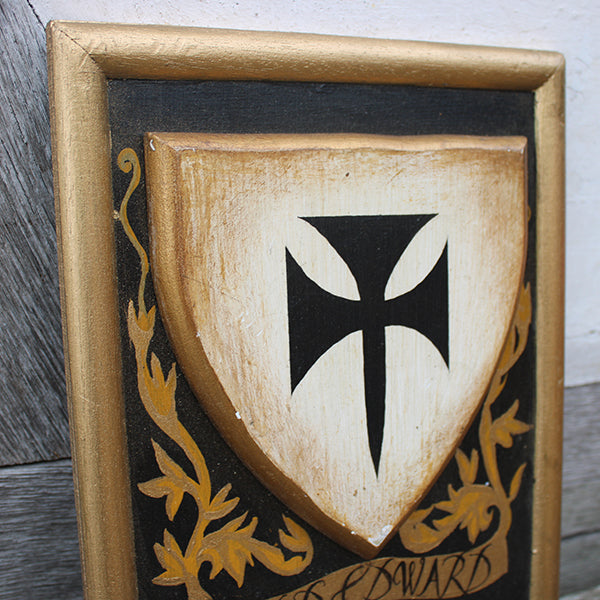 Wooden shield with black cross design and gold accents, framed in gold, on a wooden surface.