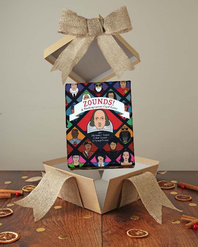 
                  
                    Black, red, pink, yellow, green and blur portrait orientated box with Shakespeare on the cover, popping out of kraft box with bow on wood base with Christmas decor
                  
                