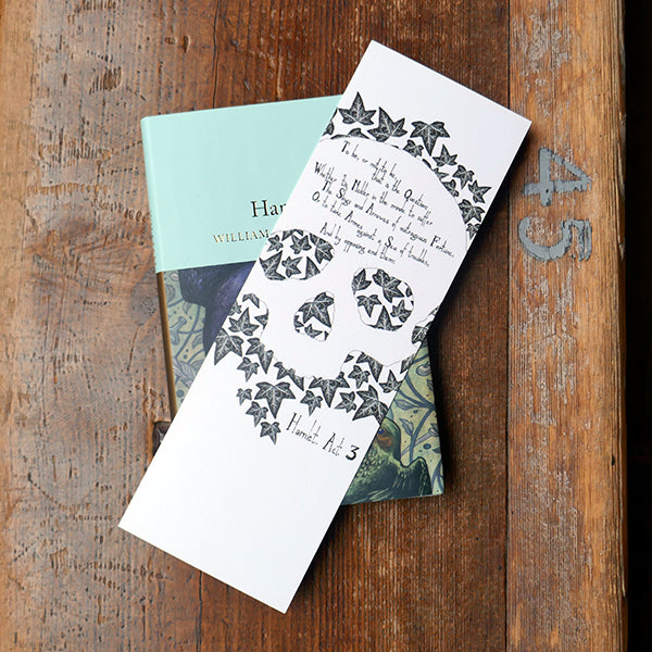 Large card bookmark with a white background, printed with a black and white image of a human skull on a bed of ivy leaves. A quote from Shakespeare play, Hamlet , "To be or not to be..." is printed over the forehead of the skull. Shown with a copy of 'Hamlet' by William Shakespeare.
