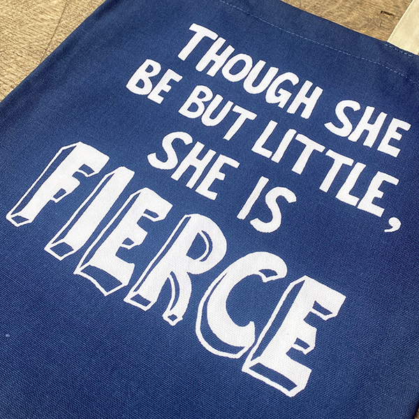 A mid-blue bag with black mid-length handles. The bag has a printed quote from Shakespeare play, A Midsummer Night's Dream (though she be but little, she is fierce.) The quote is printed in white and is made from hand-drawn capital letters