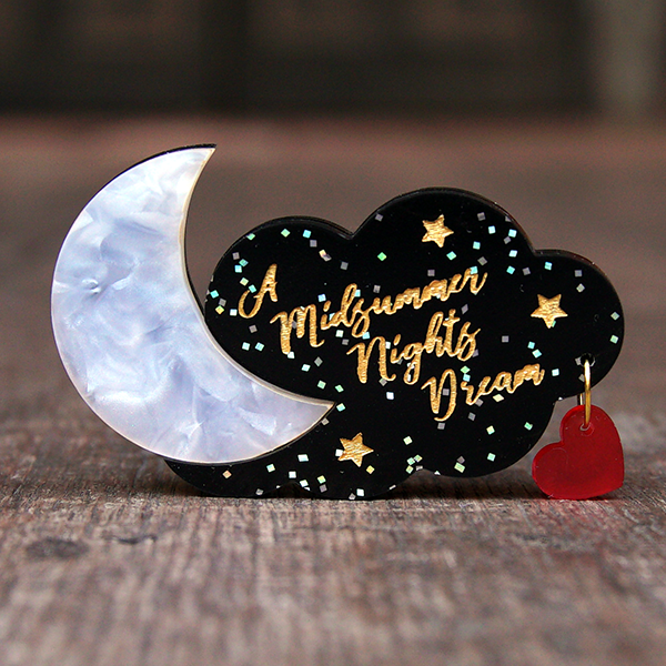 Brooch made of coloured acrylic Perspex. A black cloud with silver glitter forms the background and is engraved with the title of Shakespeare play, A Midsummer Night's Dream in gold, cursive lettering. The cloud is pierced on one side with a small metal ring. On the ring hangs a transparent red heart. On the other side of the cloud is a Perspex cresent moon which looks as though it is made of mother-of-pearl.